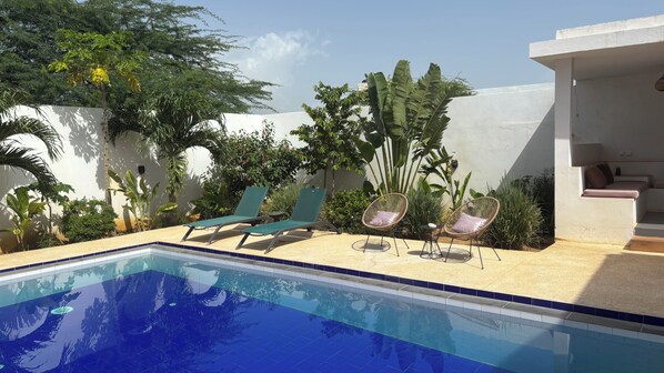 Une piscine entourée de chaises longues et d’une petite table, le tout agrémenté de plantes tropicales et bordé d’un mur blanc.