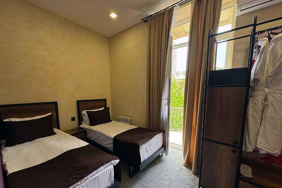 A hotel room with two single beds, a radiator, a window with curtains, and a balcony with a clothes drying rack.