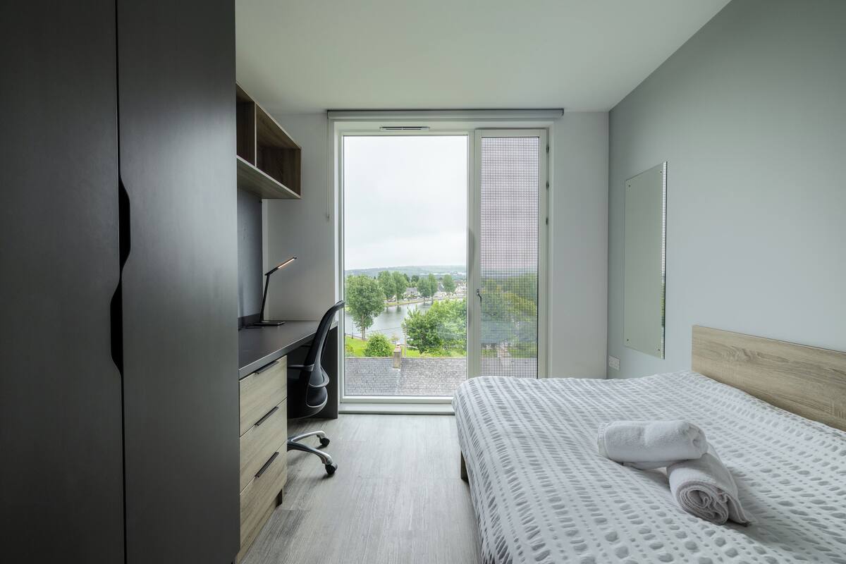 Ein modernes Hotelzimmer mit einem großen Fenster, einem Bett mit weiß gestreifter Bettdecke, einem Schreibtisch mit Stuhl und einem Kleiderschrank.