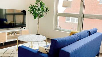En moderne stue med en blå sofa, et rundt hvidt bord, et fjernsyn på et træstel og et stort vindue med udsigt til bygninger.