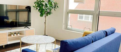 A modern living room with a blue sofa, a round white table, a TV on a wooden stand, and a large window with a view of buildings.