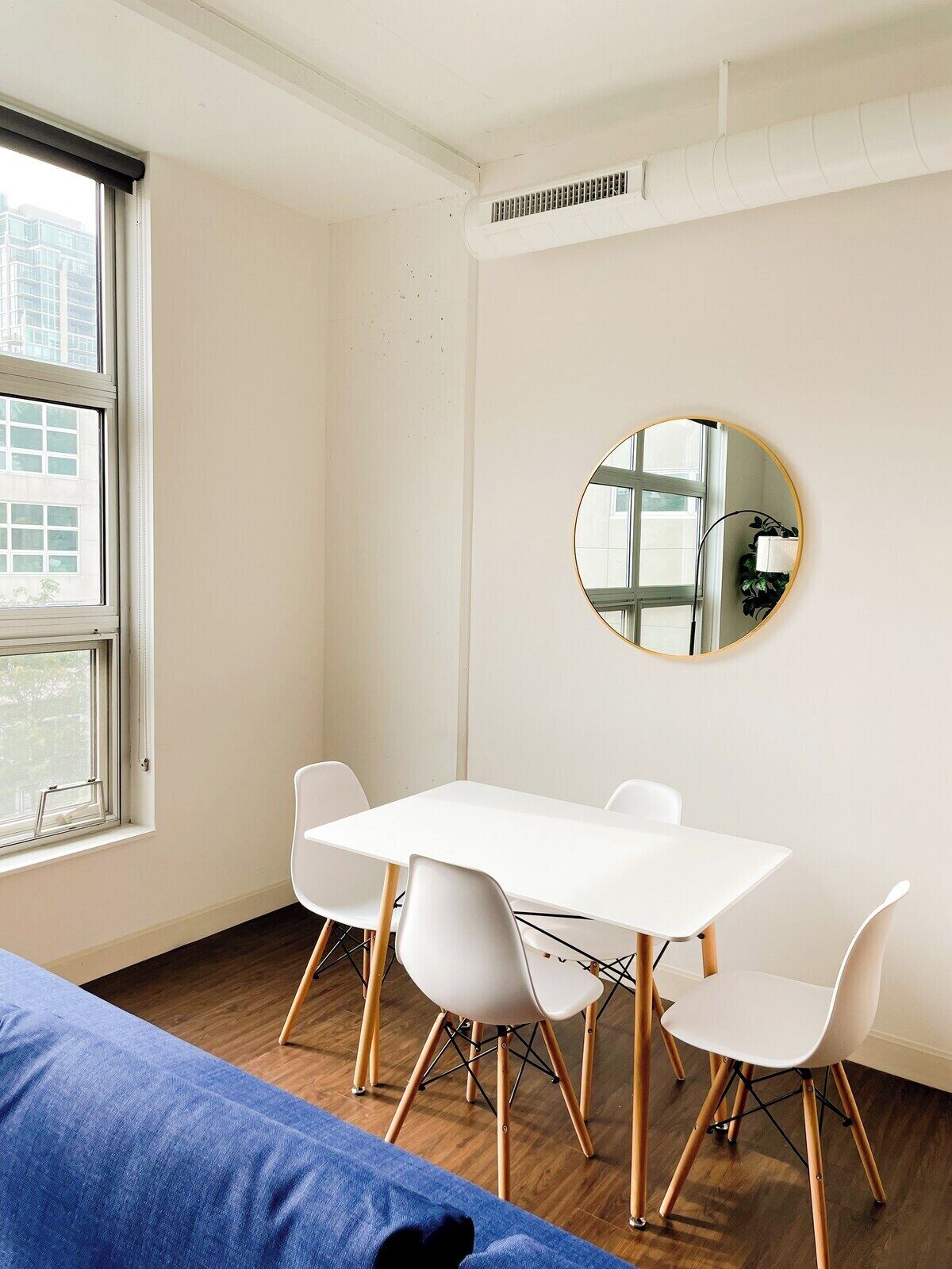 Een moderne, minimalistisch ingerichte kamer met een witte tafel en vier witte stoelen, een ronde spiegel aan de muur en een groot raam met uitzicht op gebouwen.