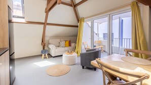 A modern living room with a sofa, ottoman, and dining table. There is a balcony with yellow curtains and a view of the outside. - Bright Historic Loft in the Heart of Willemstad (Willemstad)