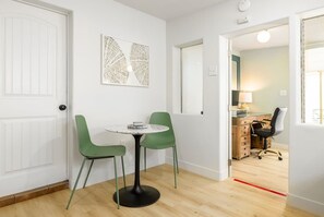 A room with a white door, a round table with two green chairs, a wooden desk with a chair, and a wall-mounted artwork.
