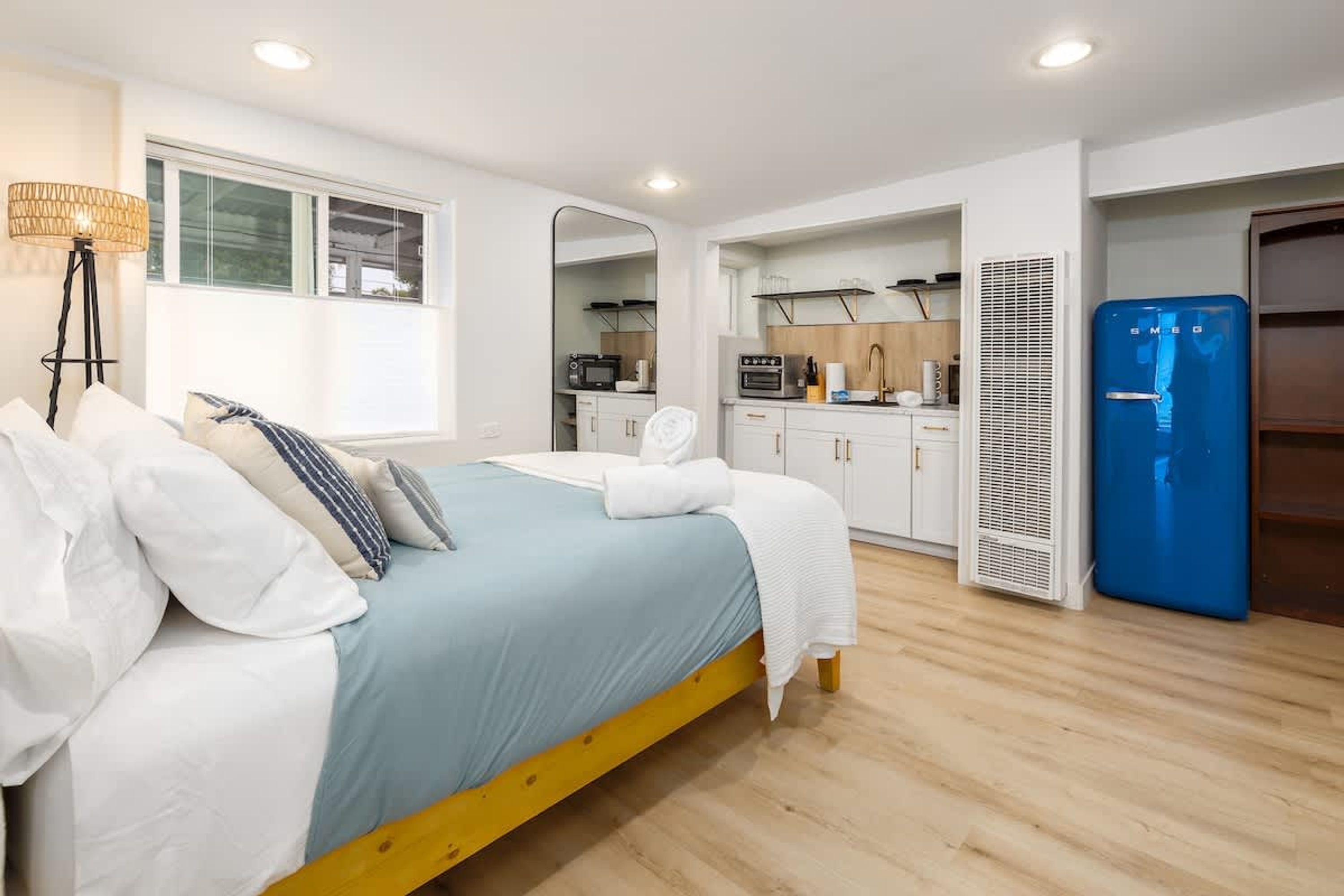 A bedroom with a bed, bedside table, lamp, and a view of a kitchenette with a microwave, toaster, and refrigerator.