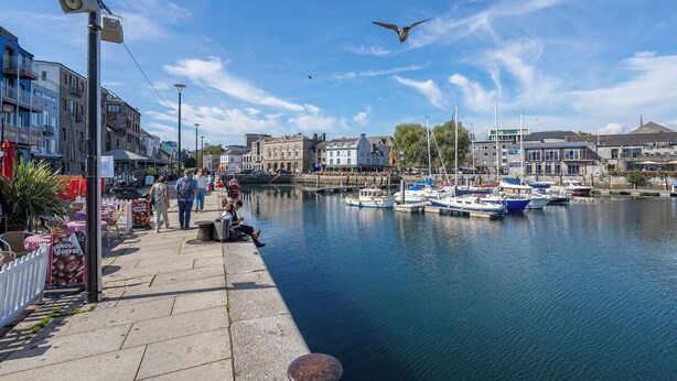 A waterfront promenade with boats docked, buildings, and a clear sky.
