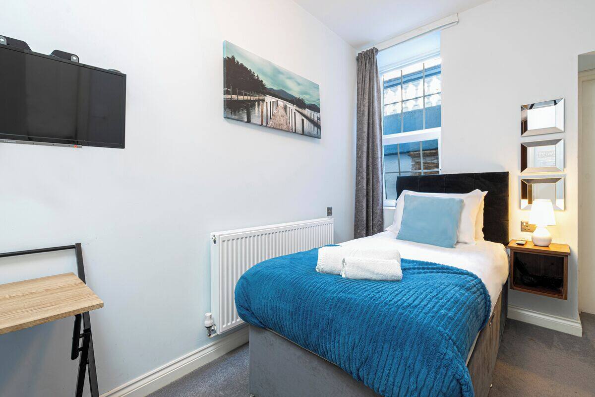 A hotel room with a bed, a nightstand, a TV mounted on the wall, a window with curtains, and a painting above the bed.