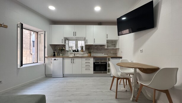 Una cocina moderna con armarios blancos, horno empotrado y comedor con mesa de madera y sillas blancas.