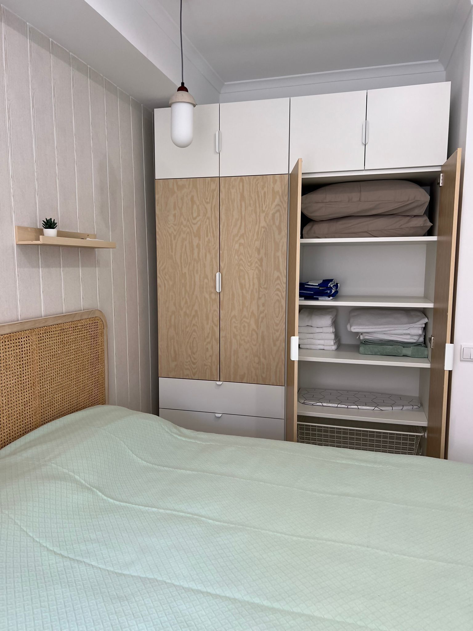 A bedroom with a bed, a wardrobe with multiple compartments, a hanging light fixture, and a small shelf with a plant.