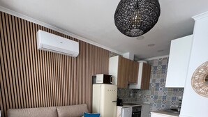 Un salon moderne avec un canapé beige, un petit pouf et un coin cuisine doté de meubles en bois et d’un dosseret carrelé.