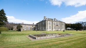 A large, historic stone building with multiple windows and a symmetrical facade, surrounded by a well-maintained lawn and a small pond. - The West Wing, Capheaton in Northumberland - sleeping 8 guests in 4 bedrooms (Capheaton)