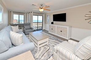A living room with a sofa, armchair, and ottoman, a ceiling fan, a TV, and a view to the outside. - Surfwatch II 103 – Stunning Oceanfront 2 Bedroom Retreat (Surfside Beach)