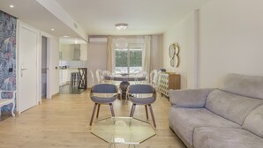 Una sala de estar con un sofá, una mesa de vidrio y dos sillas. Se observa un comedor con una mesa y sillas al fondo.