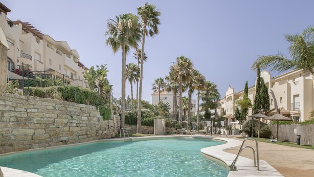 Una piscina con agua cristalina, rodeada de muros de piedra y palmeras, con edificios al fondo.