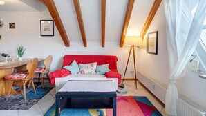 Ein Zimmer mit einer roten Couch, Holzstühlen, einem Tisch mit Lampe und einem Fenster mit weißen Vorhängen.