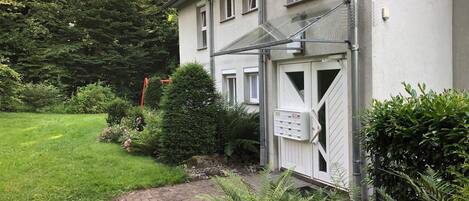 Ein Wohngebäude mit gepflasterter Auffahrt, einem befestigten Eingangstor und einem gepflegten Garten mit unterschiedlichen Pflanzen und einem kleinen Teich.