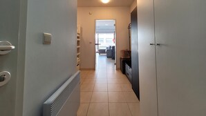 Un pasillo estrecho con un radiador, una puerta a la derecha y una vista hacia una sala de estar con un sofá y una ventana.