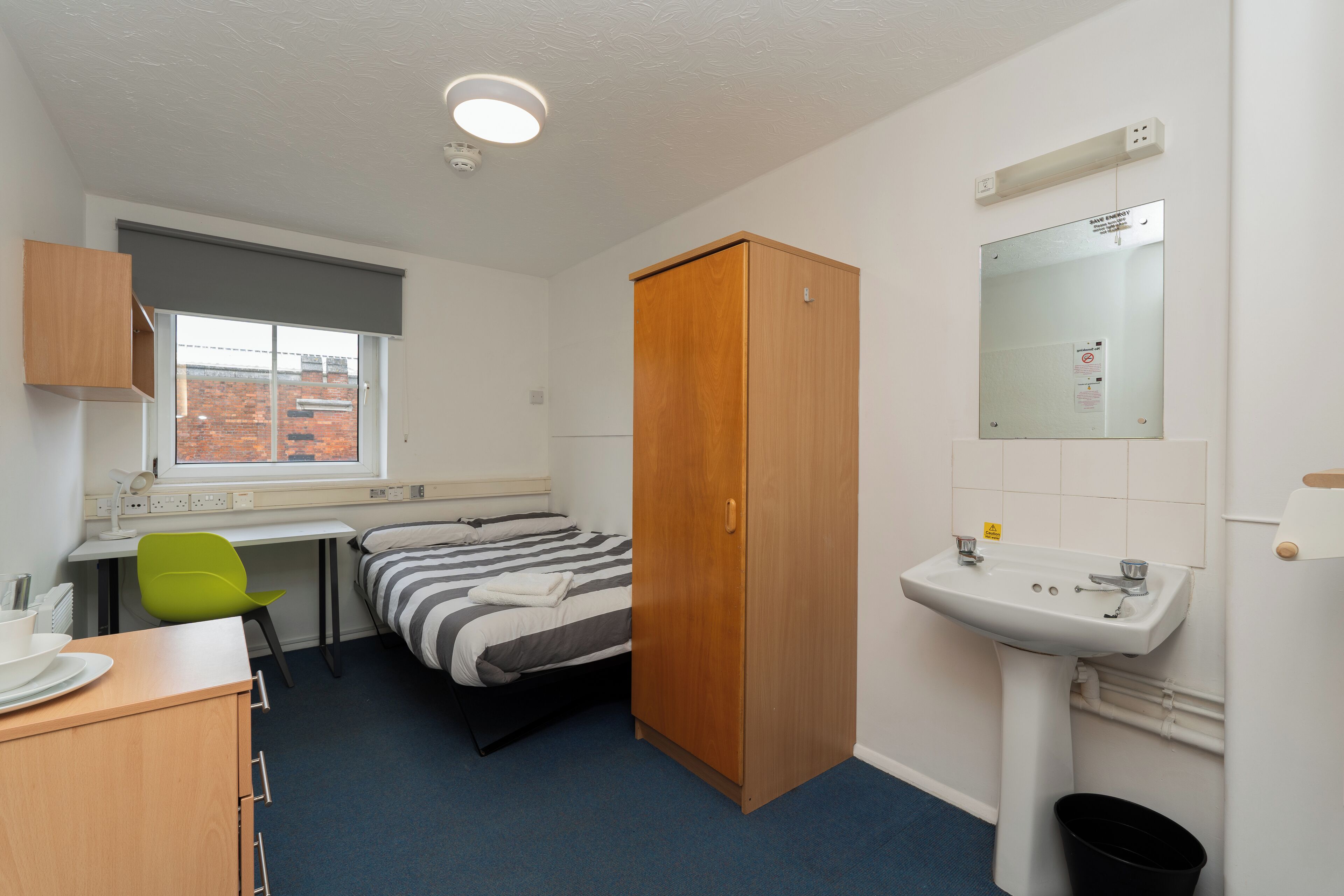A compact room with a bed, a desk, a chair, a wardrobe, a sink, and a window with a view of a brick building.