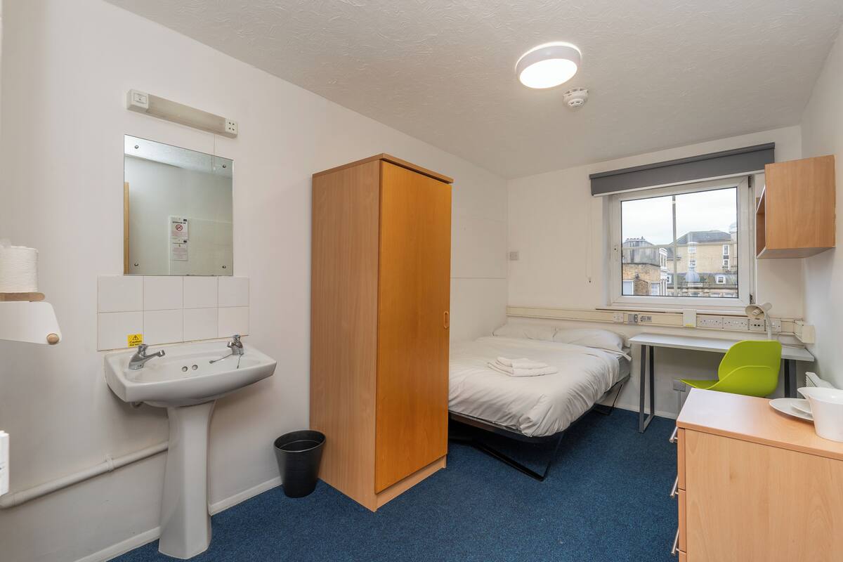 Habitación de hotel con cama, escritorio, silla, lavamanos y armario.