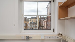 Une pièce comprenant un lit, un bureau, une fenêtre donnant sur des immeubles et des prises électriques au mur.