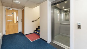 Un couloir avec une moquette bleue, une porte en bois portant l’inscription « Appartement 3A », un tapis rouge au pied d’un escalier et un ascenseur.