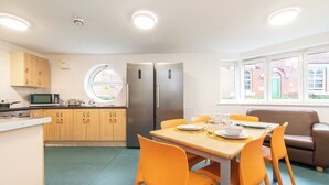 Une cuisine compacte équipée d’un réfrigérateur, d’un four à micro-ondes et d’une cuisinière, une table à manger avec des chaises orange et une fenêtre donnant sur des immeubles.