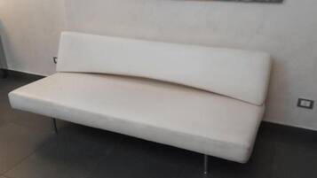 Un sofá blanco con patas metálicas en una habitación con suelo de baldosas oscuras y una pared lisa.