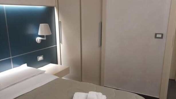 A neatly made bed with a folded towel, a bedside lamp, and a wall-mounted light fixture in a modern hotel room. - FULL HOME. (Cosenza)