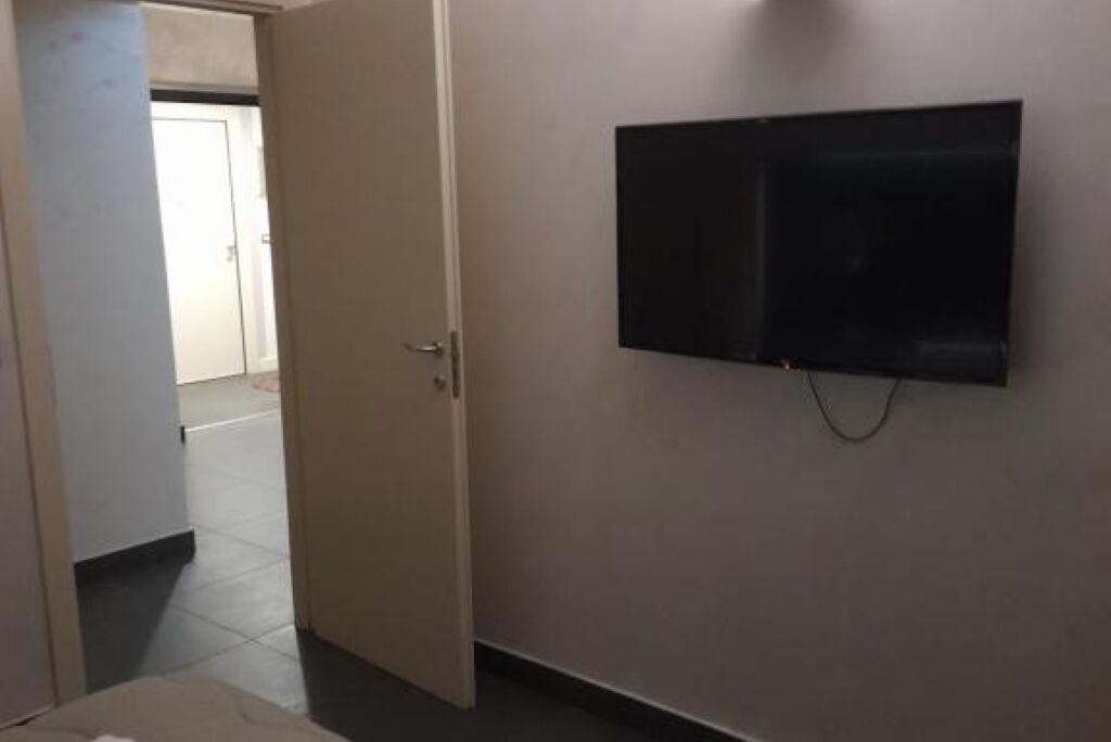 A hallway with a closed door, a flat-screen TV mounted on the wall, and a tiled floor.