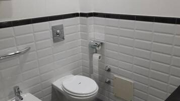 Un baño pequeño y limpio con azulejos blancos, un inodoro, un bidé y un perchero para toallas.