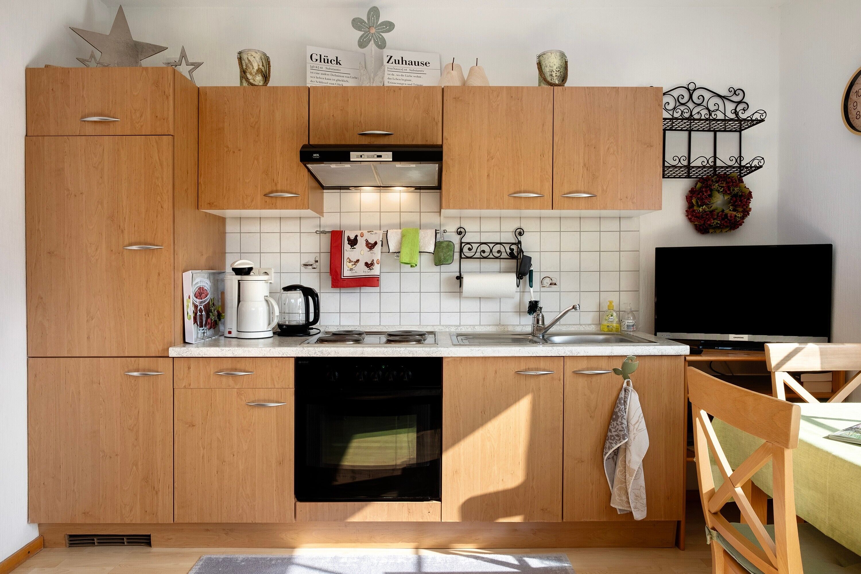 A modern kitchen with wooden cabinets, a built-in oven, a coffee maker, and a TV.
