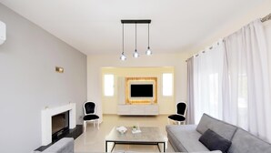 A modern living room with a grey sofa, a coffee table, and a flat-screen TV mounted above a fireplace. There are two black chairs and a large window with white curtains. - Seraina House (94QJ+W7M Pastida, Greece)
