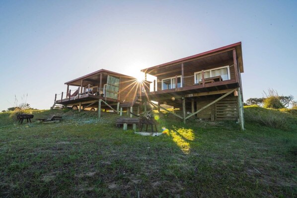 Two wooden cabins with elevated platforms, situated on a grassy hill with a clear sky.