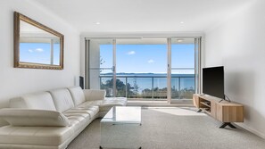 A modern living room with a sofa, a coffee table, a TV on a wooden stand, and a large sliding glass door leading to a balcony with a view of the sea.