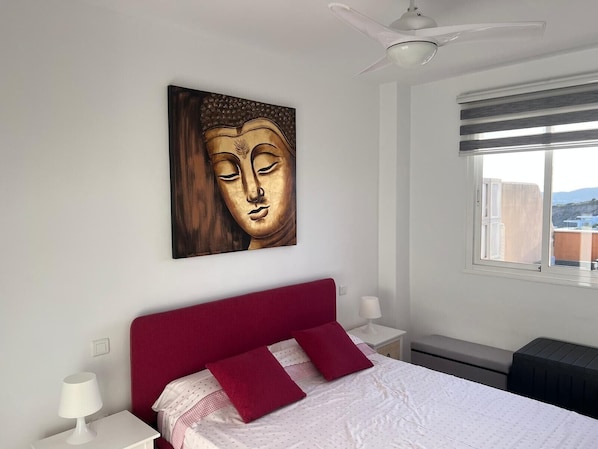 Une chambre à coucher comprenant un lit, un ventilateur de plafond, une fenêtre avec vue et une œuvre d’art accrochée au mur.