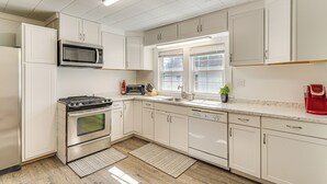 Una cucina moderna con mobili bianchi, elettrodomestici in acciaio inossidabile, forno a microonde, fornello e lavastoviglie. Una finestra lascia entrare luce naturale e un piccolo vaso con una pianta è posizionato sul piano di lavoro.