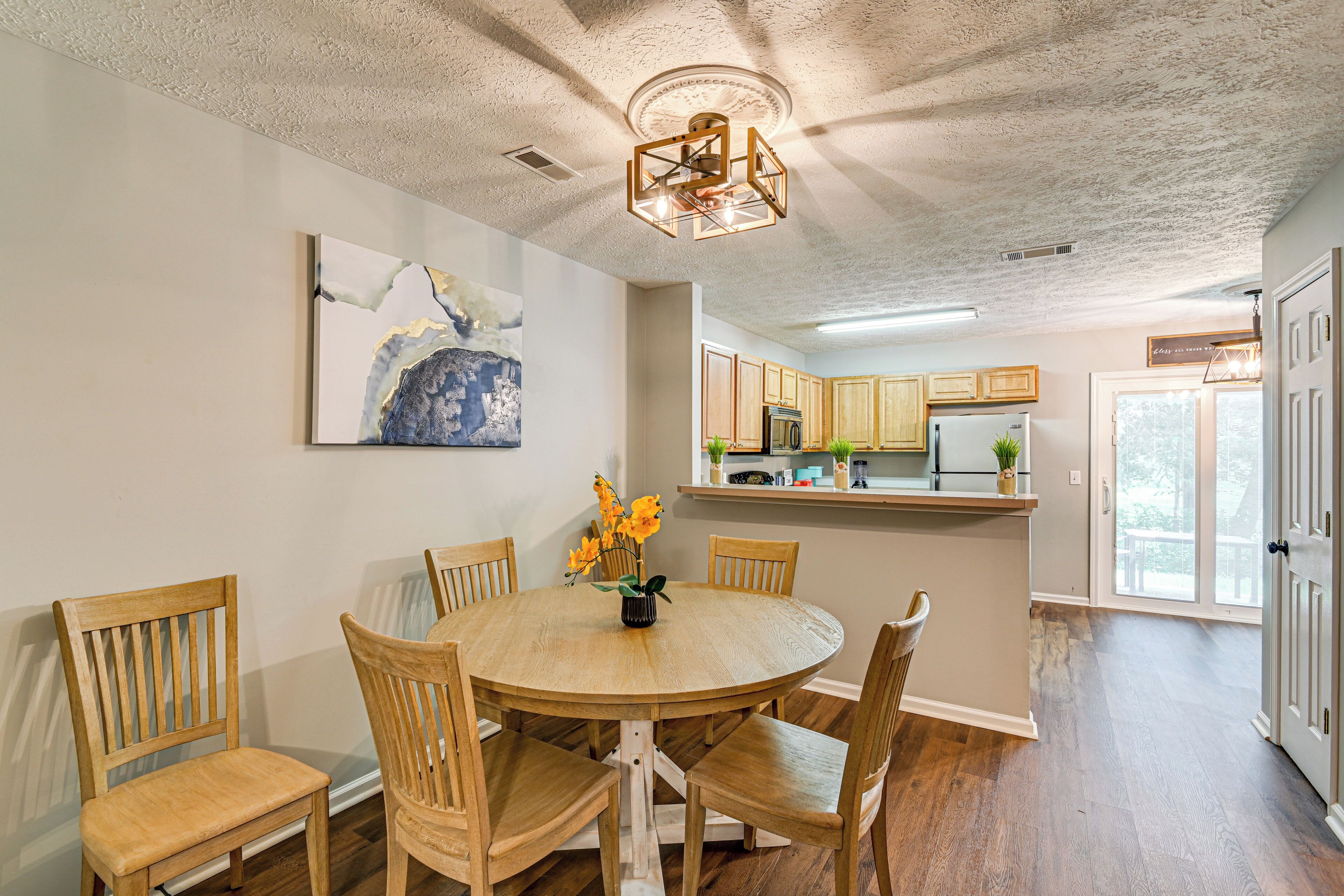 A dining area with a round wooden table, six chairs, a ceiling light fixture, and a painting on the wall.
