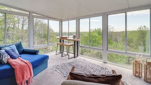 Et værelse med en blå sofa, et træbord og udsigt til grøn natur gennem store vinduer.