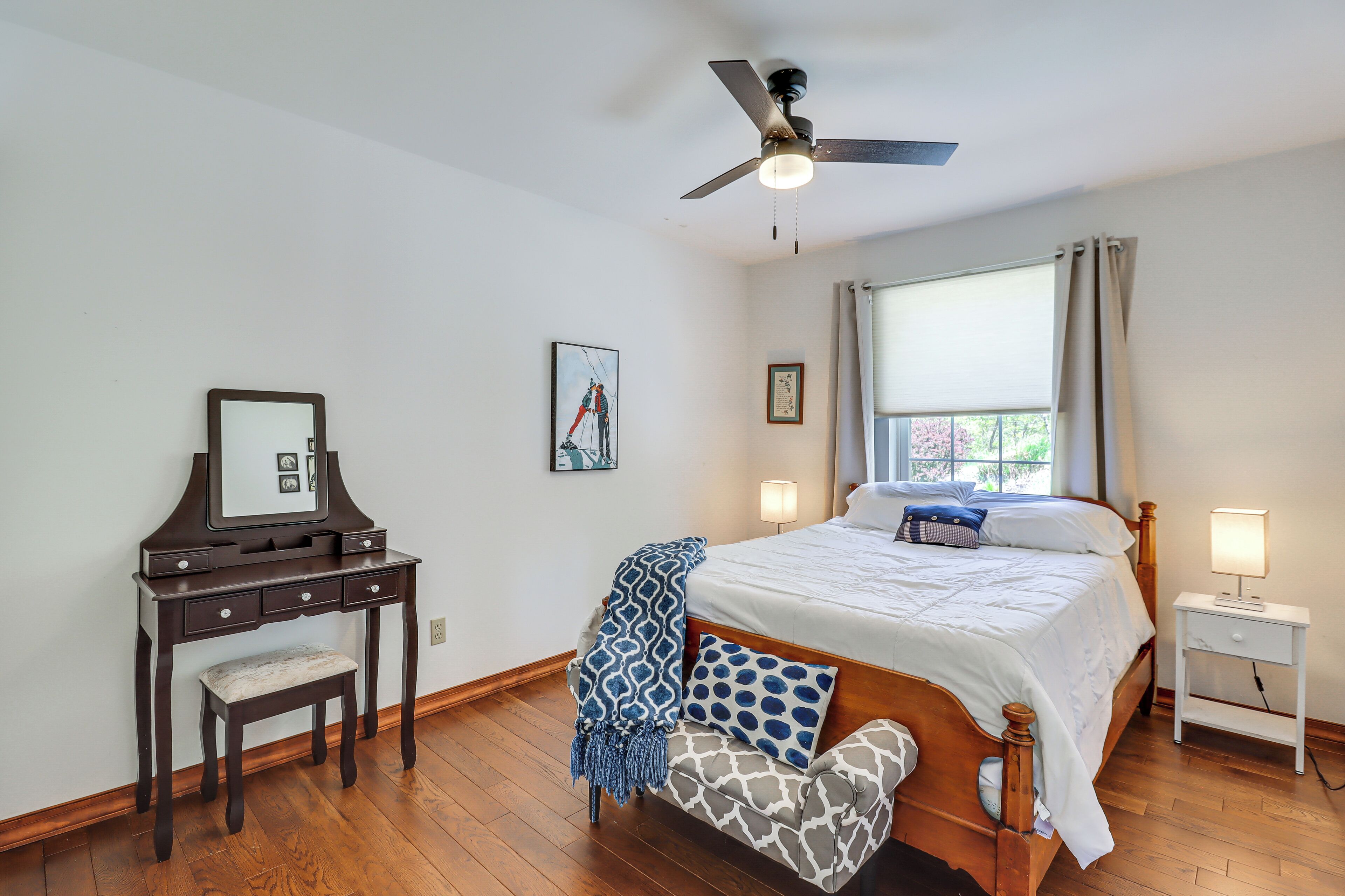 A bedroom with a bed, bedside table, nightstand, vanity with mirror, chair, and ceiling fan.