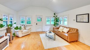 A spacious living room with a high ceiling, wooden flooring, a white sofa, a leather sofa, and a coffee table. There are large windows with curtains, a potted plant, and a framed map on the wall. - Walk to Kelsey Lake: Cassopolis Home w/ Yard! (Cassopolis)