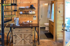 A compact kitchen with a coffee bar, wooden shelves, and a view into the adjacent bathroom.
