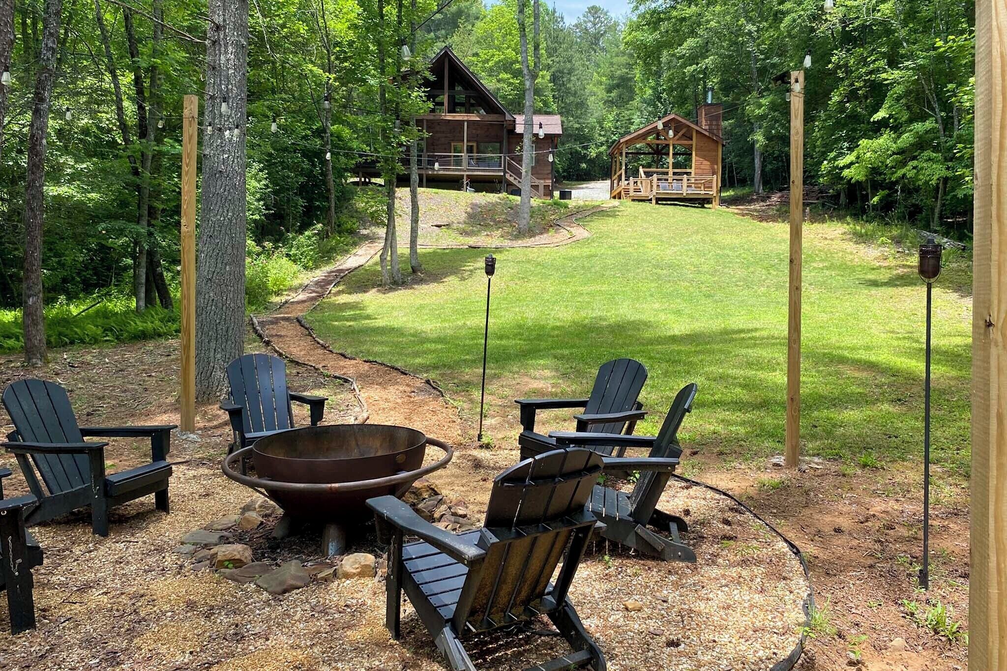 A cabin with a deck, surrounded by a forest. There are Adirondack chairs and a fire pit in the foreground.