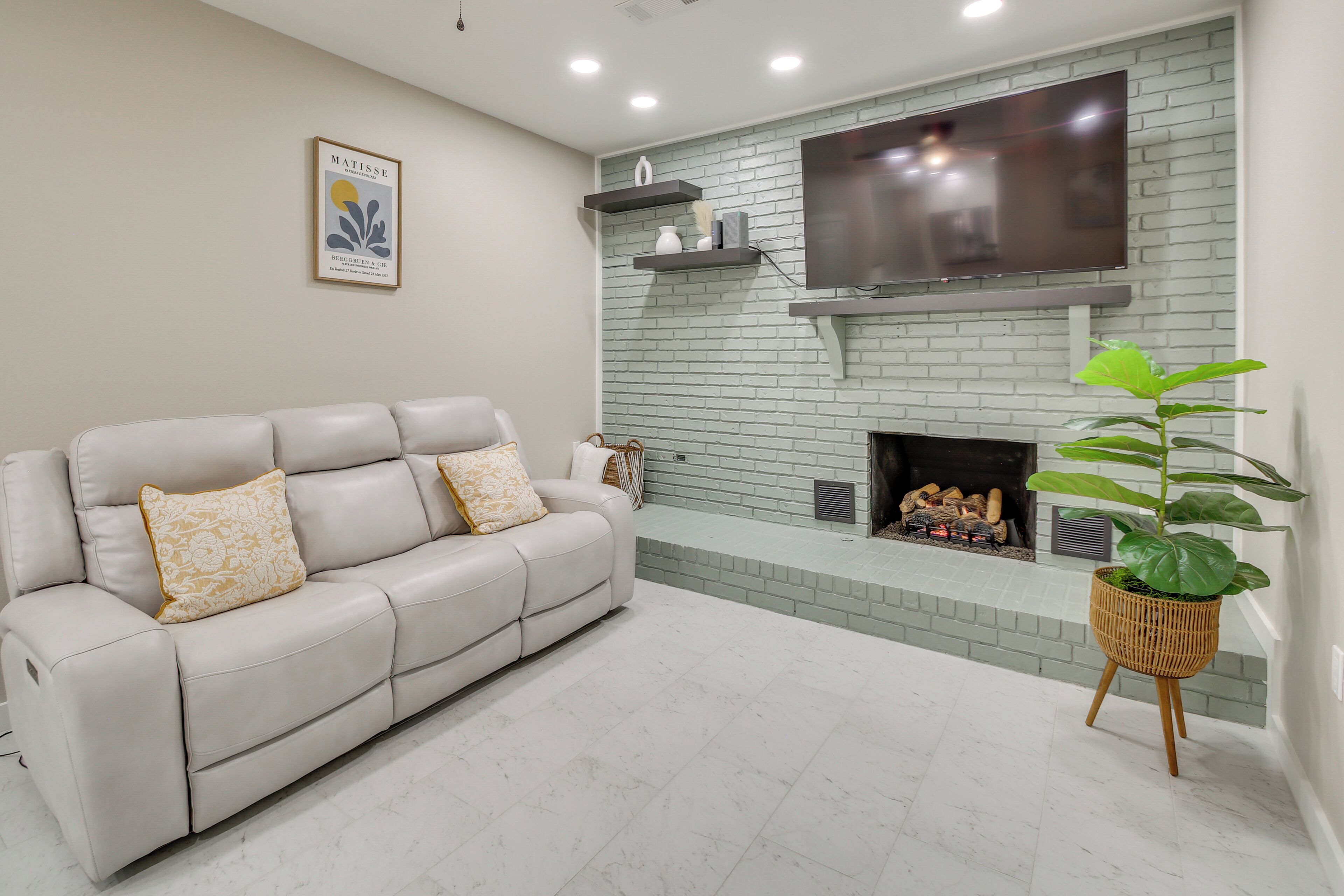 Un salon avec un canapé en cuir blanc, un téléviseur à écran plat fixé à un mur en briques, une cheminée et une plante en pot.