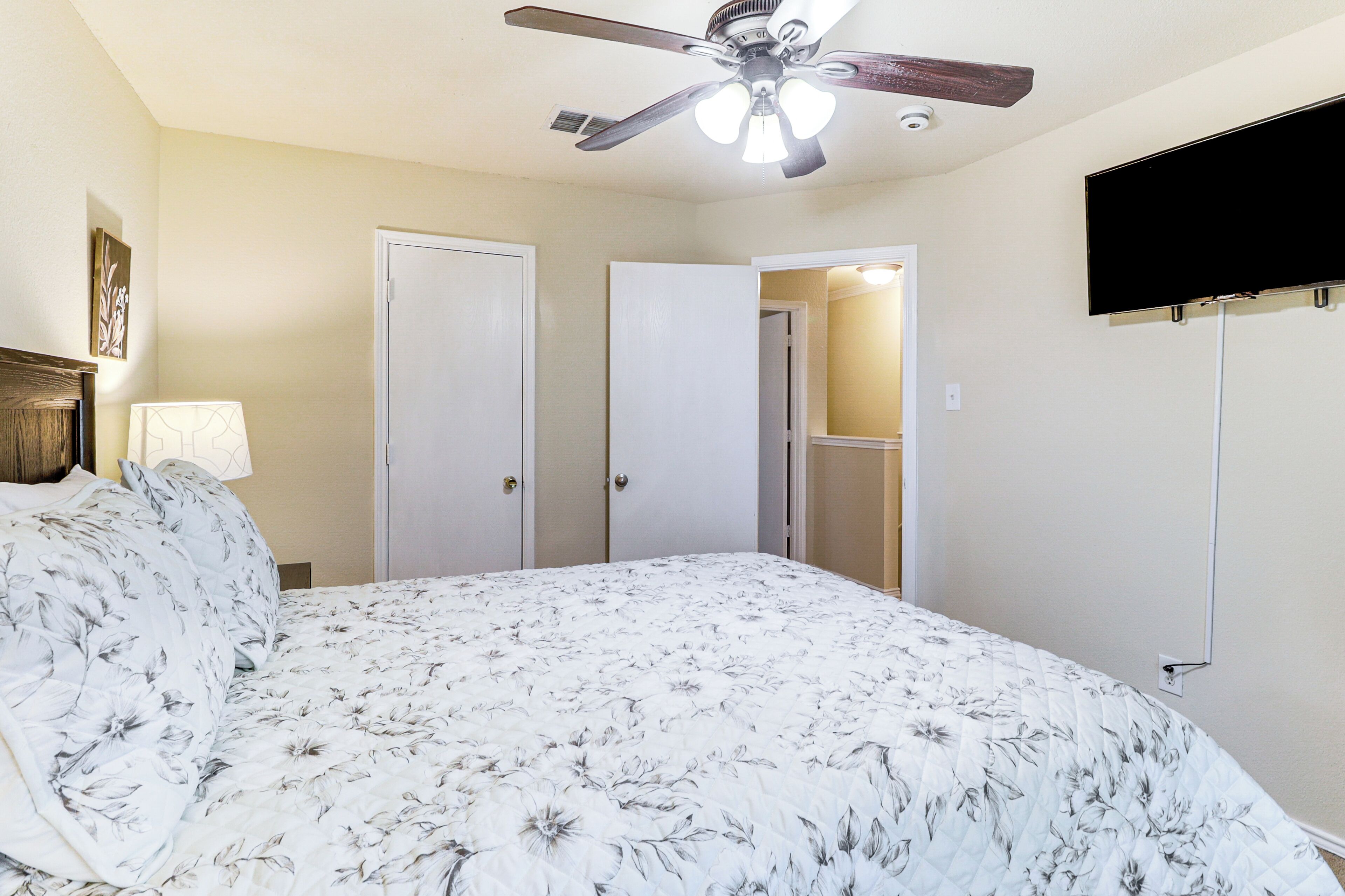 A bedroom with a bed, a ceiling fan, a TV mounted on the wall, and a door leading to another room.