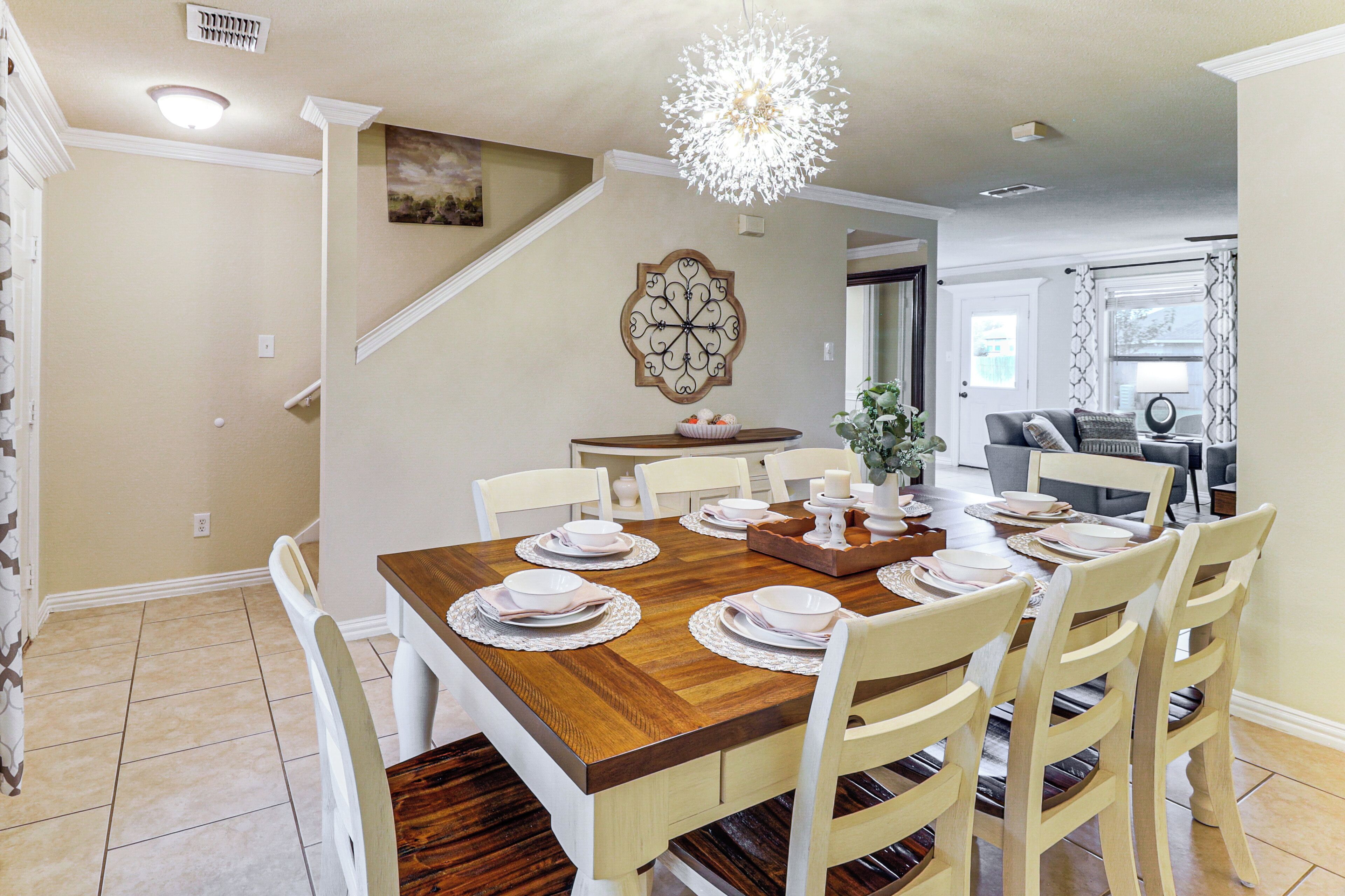 A dining area with a wooden table set for six, surrounded by white chairs. A decorative wall piece and a chandelier are visible.