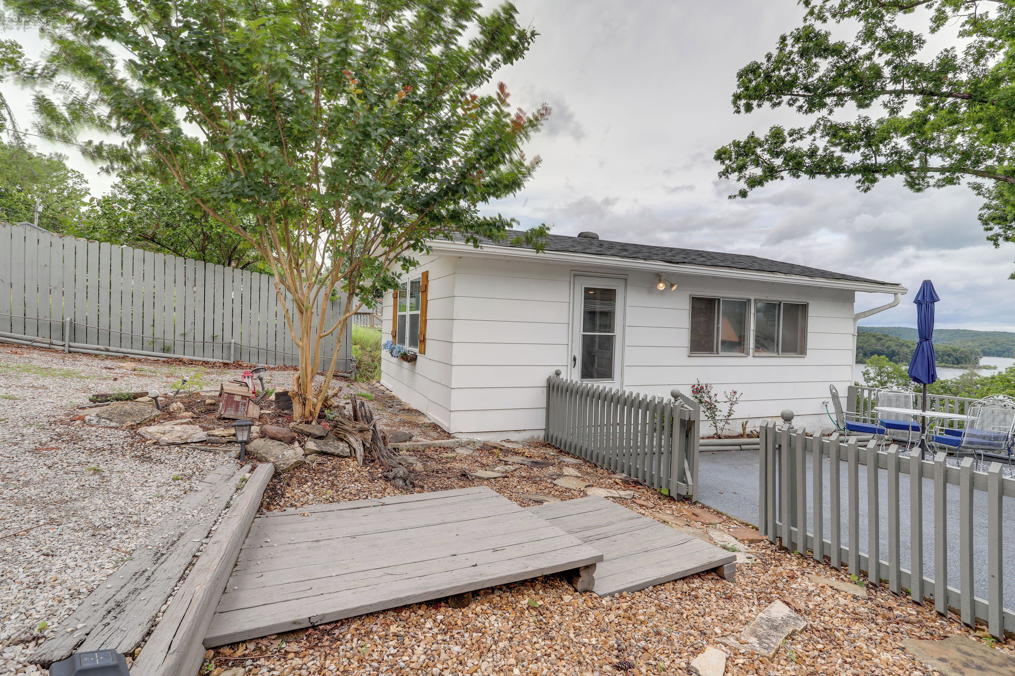 A small white house with a wooden deck, a fenced yard, and a tree.