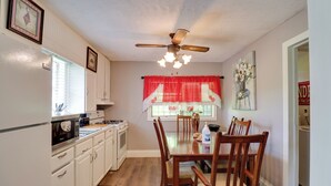 A kitchen with white cabinets, a wooden dining table, and chairs. There is a ceiling fan, a window with red curtains, and a painting on the wall. - 16 Mi to Dtwn Jackson: Quiet Southern Escape (Terry)