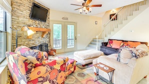A living room with a stone wall, a fireplace, a ceiling fan, and a staircase. - Pickwick Lake Cabin w/ Private Marina Access (Iuka)