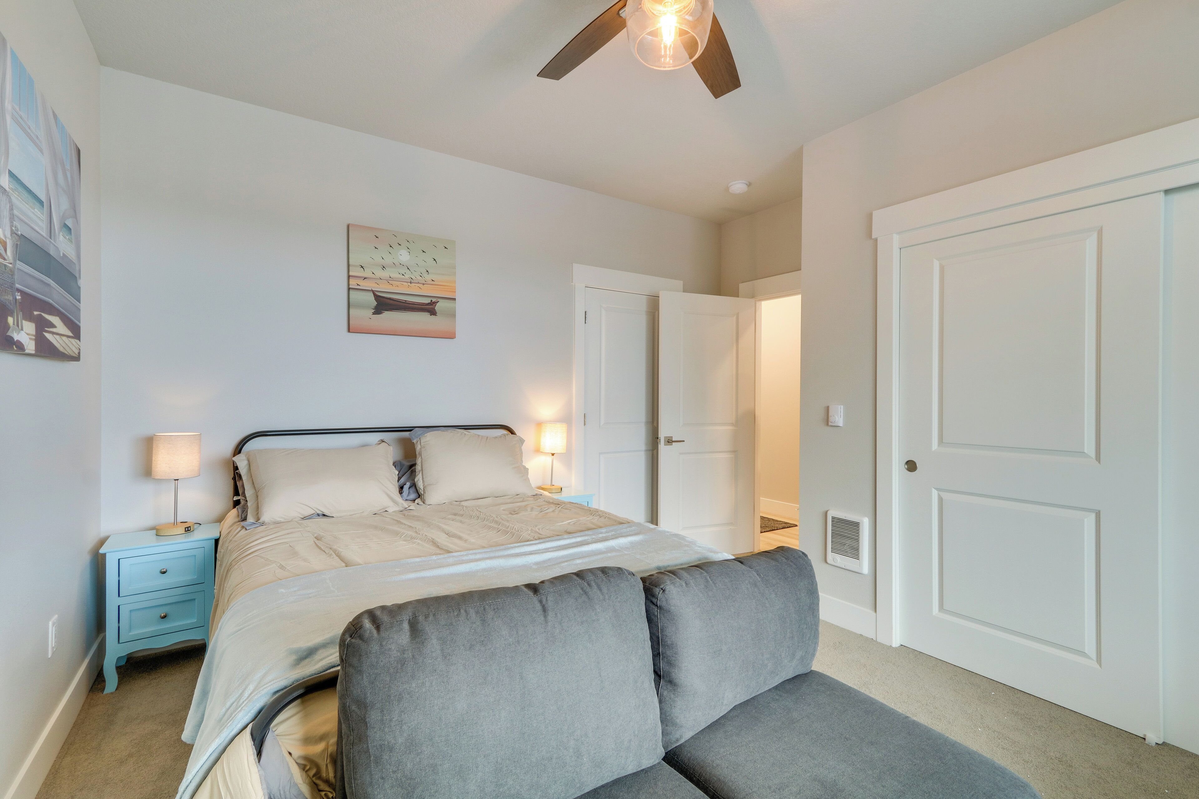 Une chambre à coucher comprenant un lit, une table de chevet, un ventilateur de plafond, une porte et un mur orné d’un tableau.