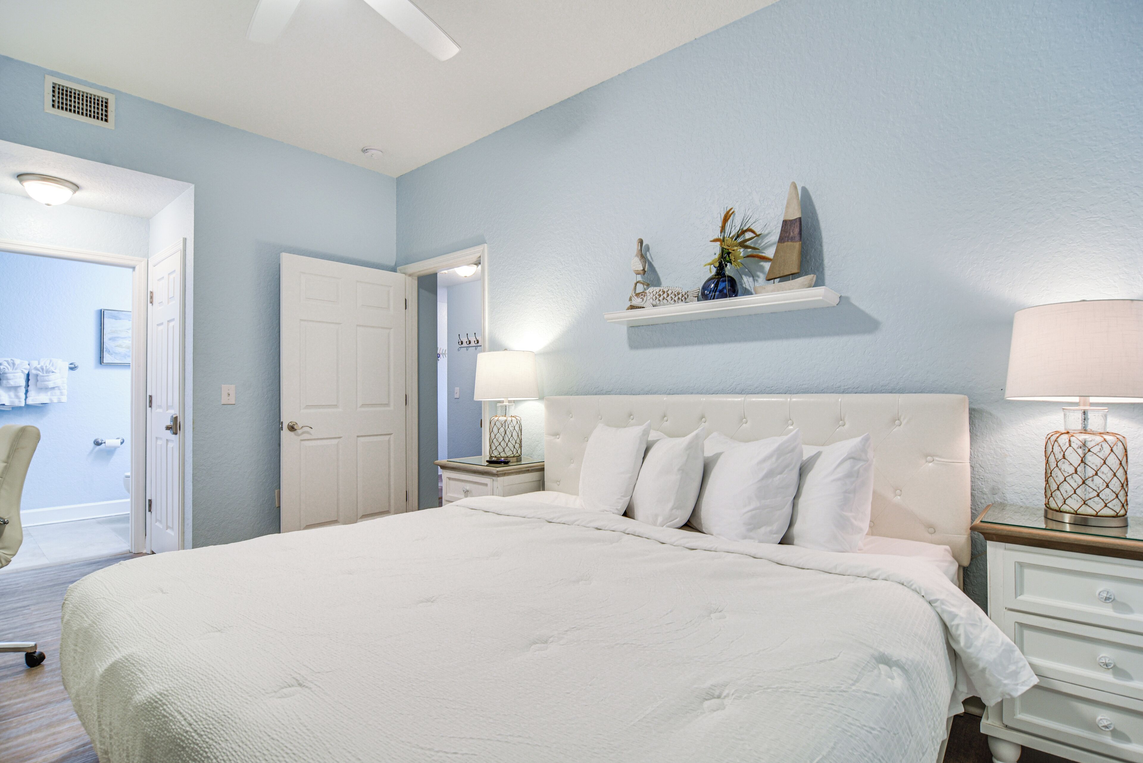 Une chambre à coucher avec un grand lit, une tête de lit blanche, des tables de chevet avec des lampes et une porte donnant sur une autre pièce.